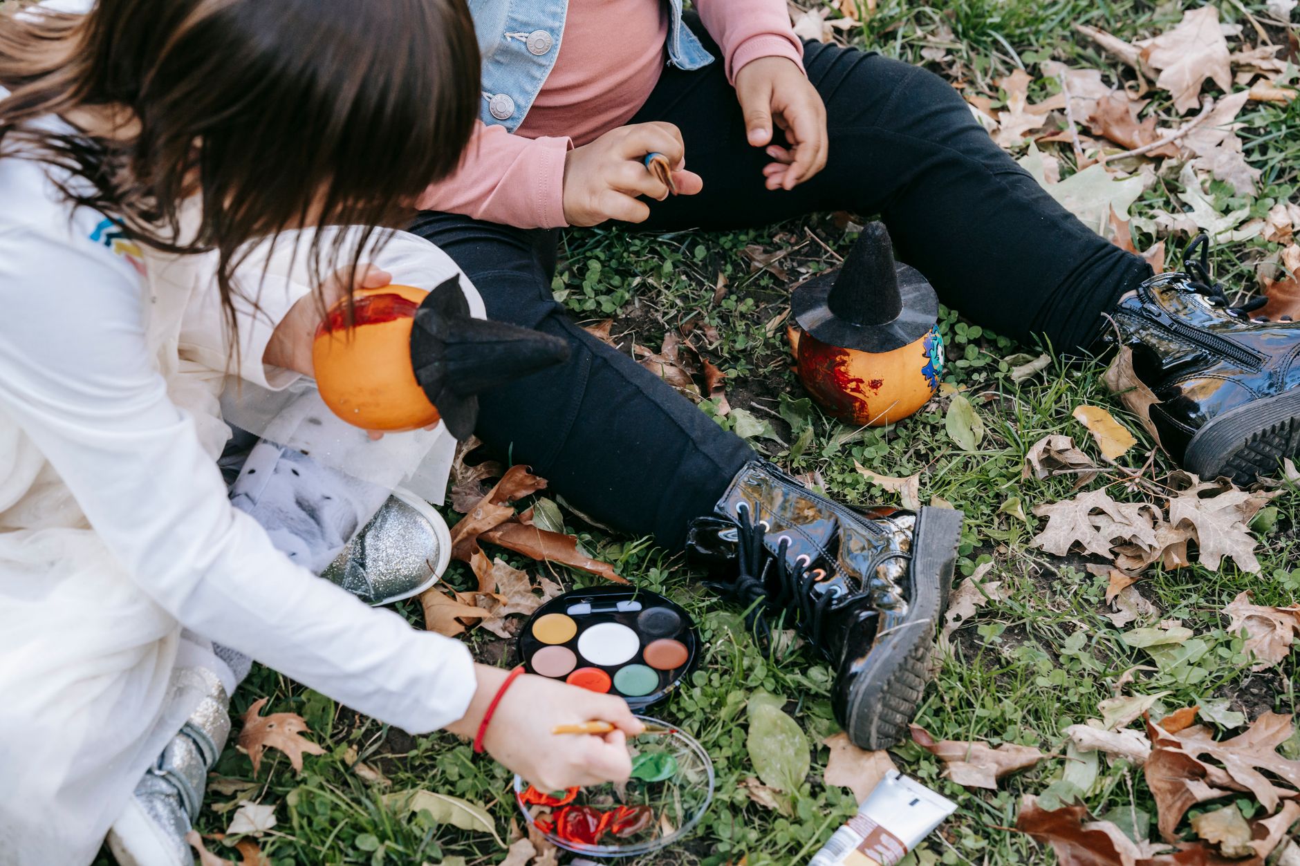 Deux enfants assis dans l’herbe parmi les feuilles mortes peignent des citrouilles, dont une coiffée d’un chapeau de sorcière, avec une palette de peinture.
