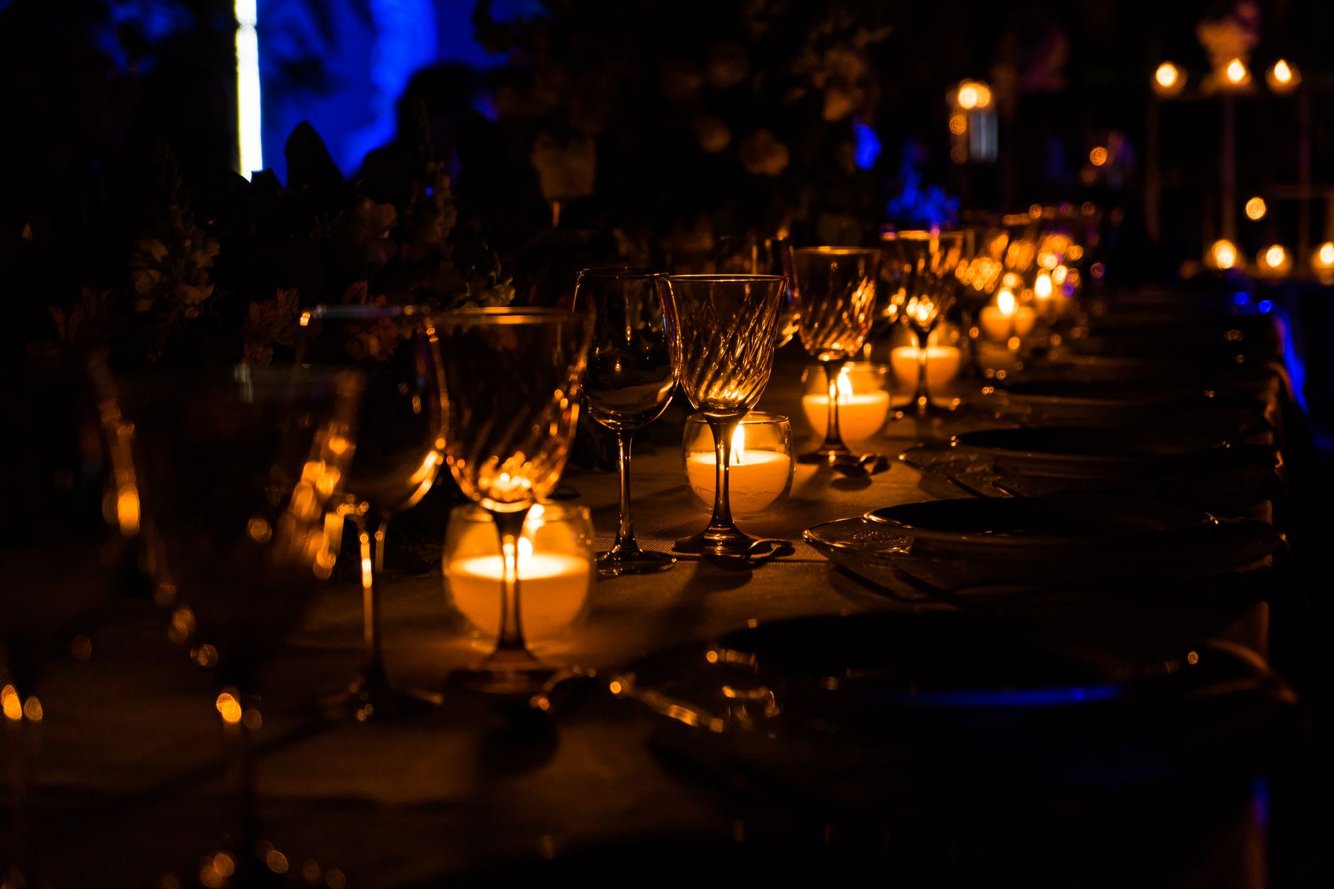 Longue table dressée dans la pénombre, avec verres à pied, assiettes et bougies allumées qui éclairent la nappe.