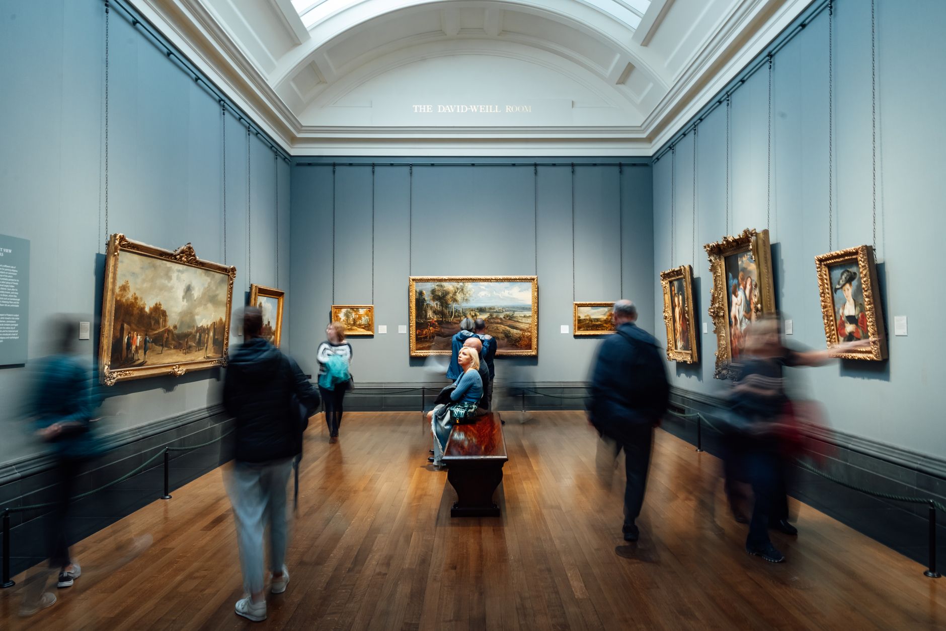 Visiteurs dans une salle de musée aux murs bleu-gris, tableaux à cadres dorés, banc central et parquet en bois.
